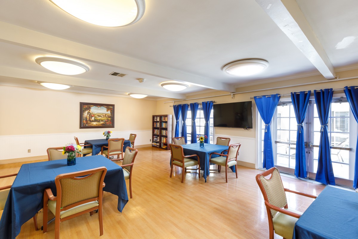 A well-lit dining area with tables and chairs, featuring blue curtains and a television on the wall.