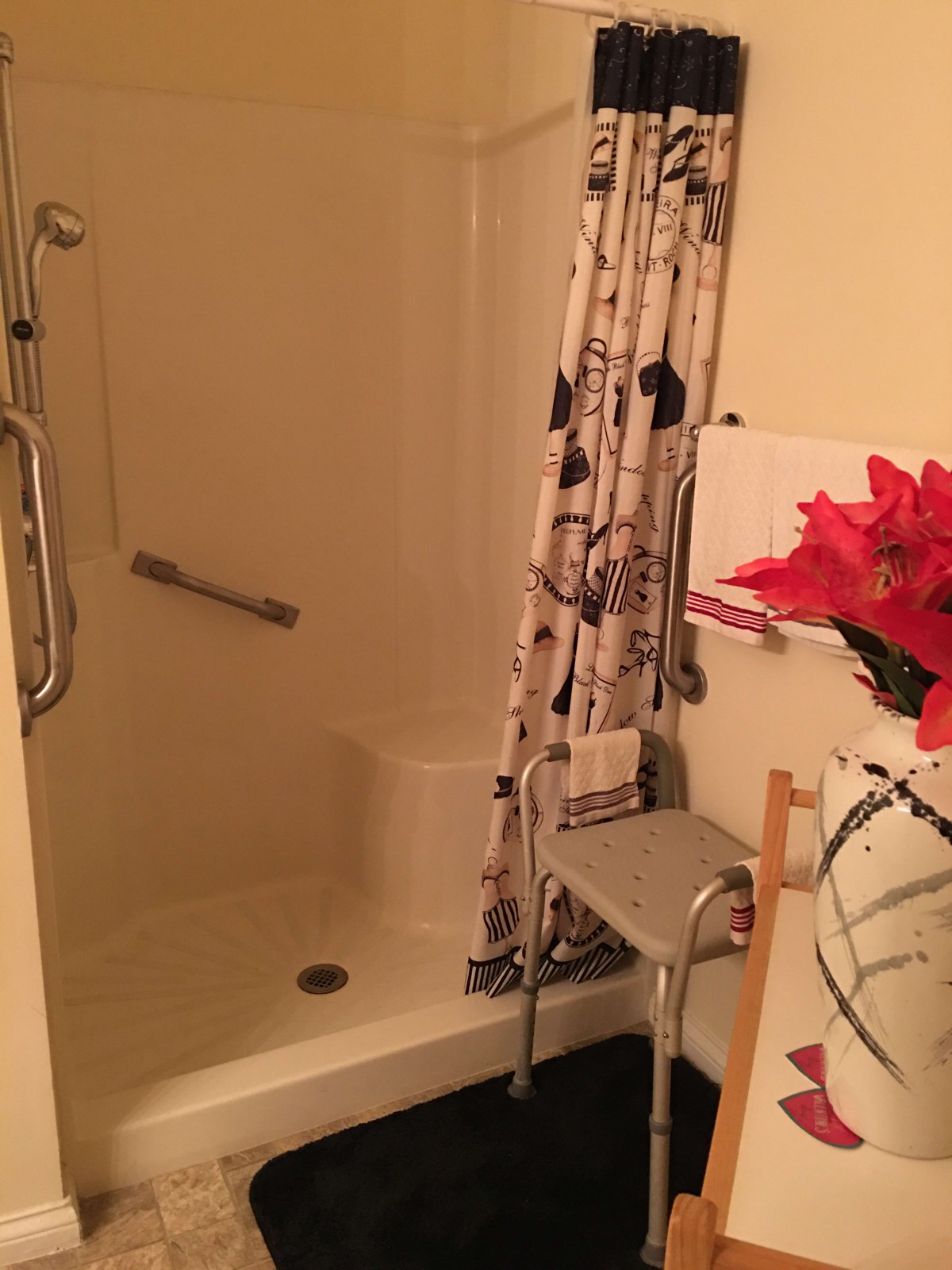 A bathroom with a shower, featuring a floral curtain and safety grab bar, with decorative flowers on the side.