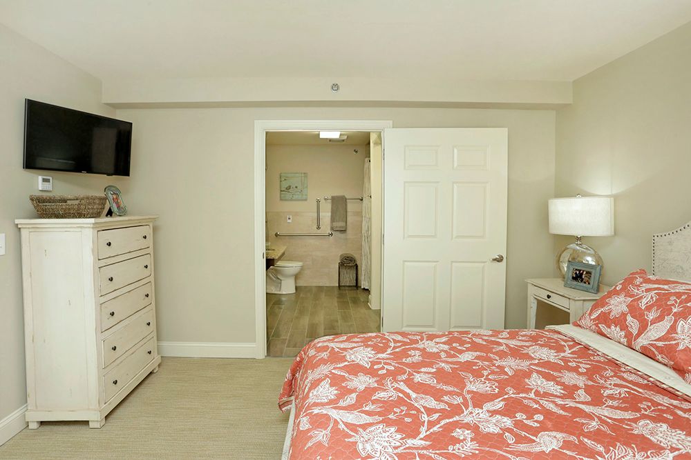 A cozy bedroom with a coral patterned bedspread, featuring a dresser and TV. The room opens to an adjoining bathroom.