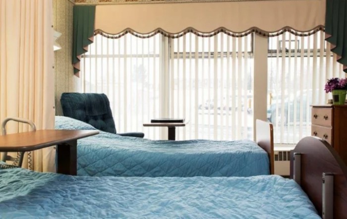 A bright bedroom featuring two neatly made beds with blue bedding and large windows letting in natural light.