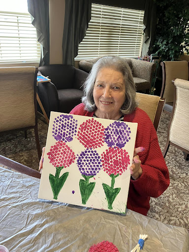 An elderly woman proudly displays her colorful painting of flowers.