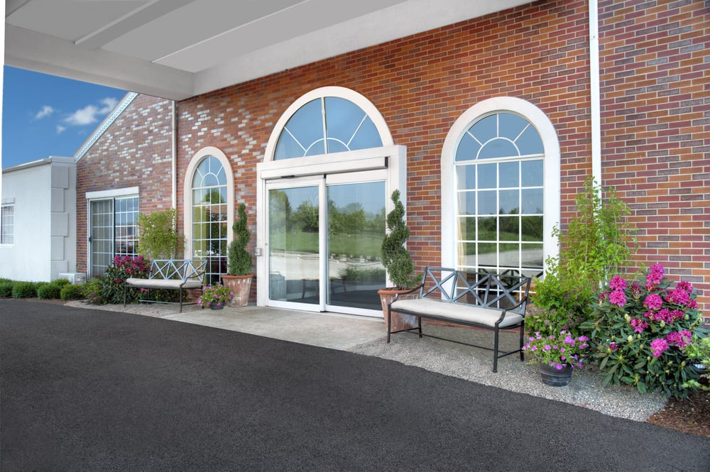 Entrance to a brick building with arched windows and glass doors, featuring benches and potted plants.