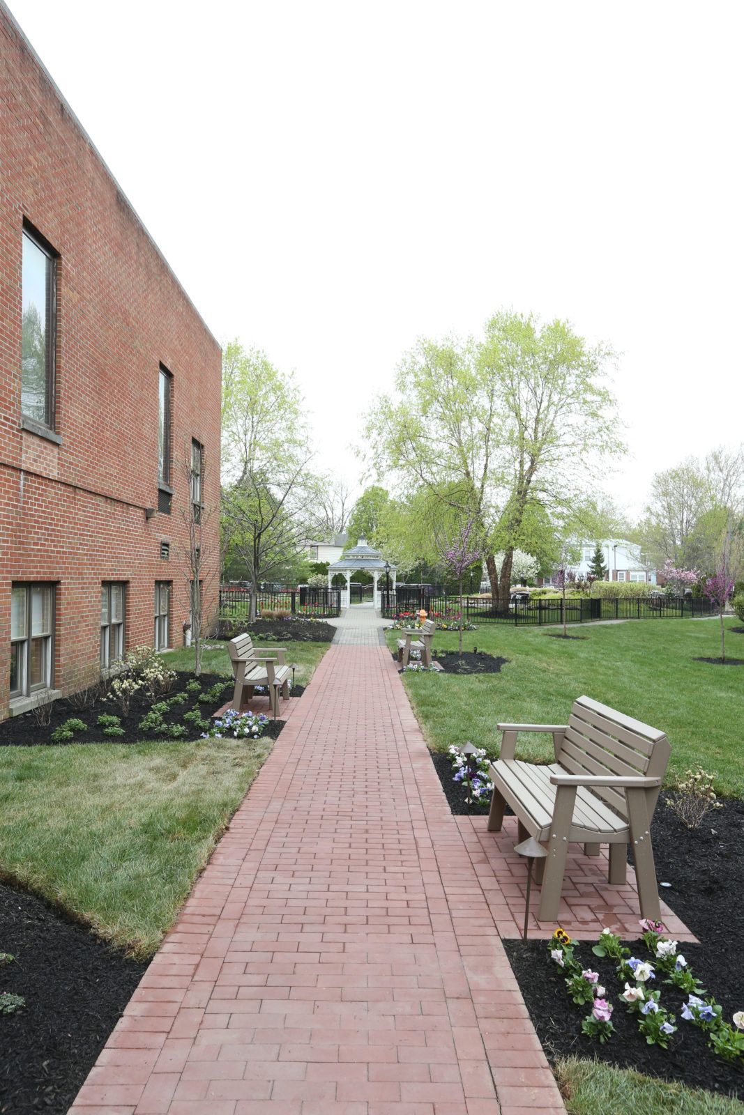 A serene outdoor space with a brick pathway flanked by benches, leading to a garden area surrounded by lush trees and greenery.