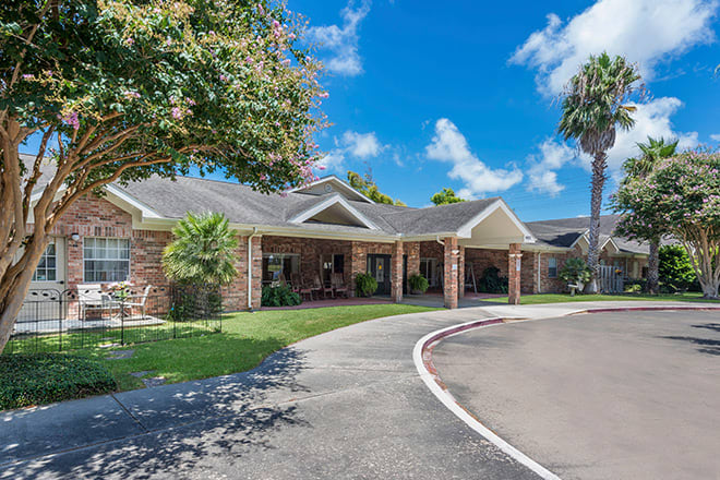A single-story brick house with a driveway and lush greenery under a clear blue sky.