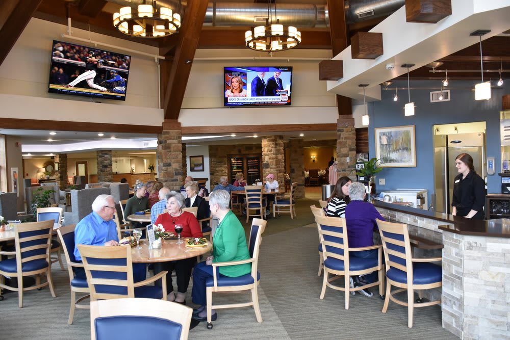 A group of seniors enjoying a meal in a cozy dining area with televisions mounted on the walls.