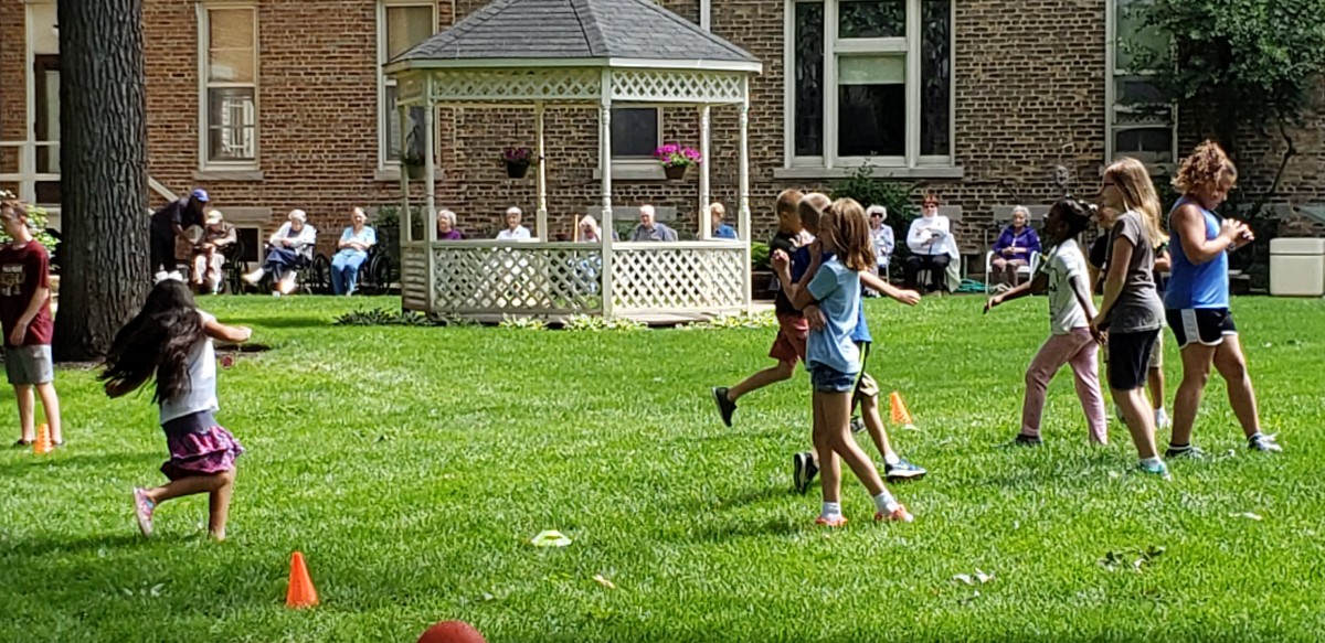 Children playing on a grassy lawn in front of a gazebo, with a group of people seated in the background.