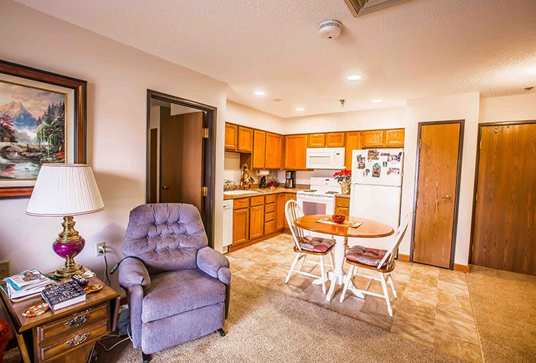 A cozy living room and kitchen area featuring a plush chair, dining table, and wooden cabinetry.