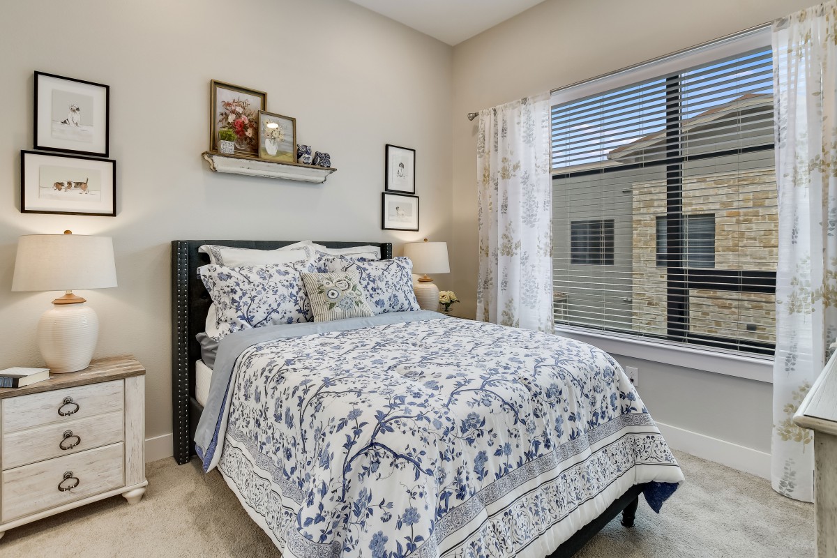 A cozy bedroom featuring a neatly made bed with patterned bedding, decorated walls with art, and a window letting in natural light.