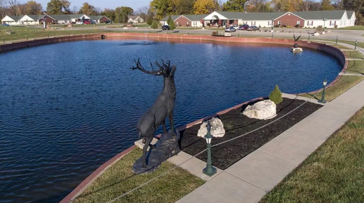 A statue of a deer stands beside a lake in a park setting.