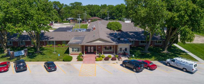 Aerial view of a building with a parking lot and cars surrounded by trees on a sunny day.