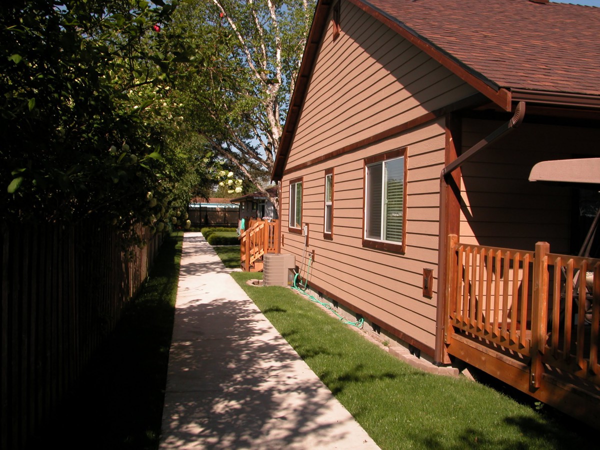 A pathway beside a house with a wooden fence and green lawn under the sunlight.