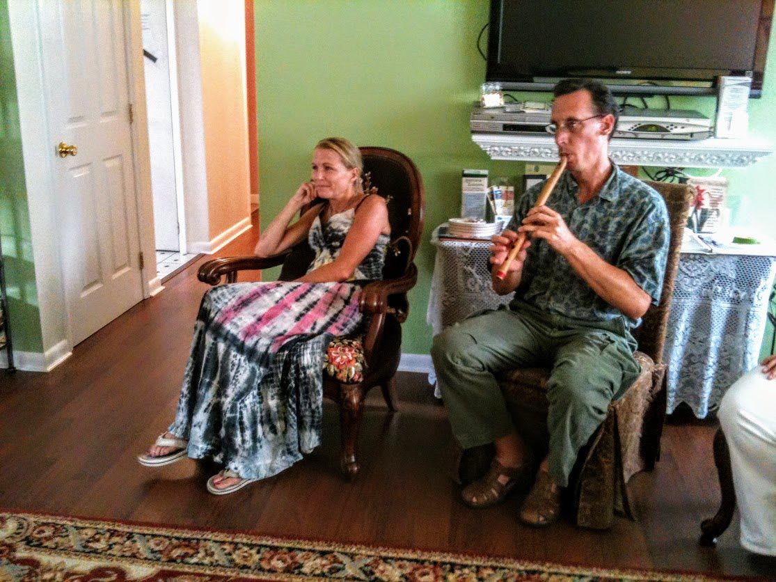 A man plays the flute while a woman listens in a comfortable living room setting.