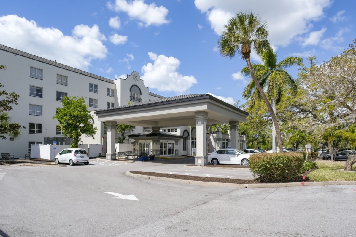 A hotel entrance with a covered driveway and surrounding palm trees under a partly cloudy sky.