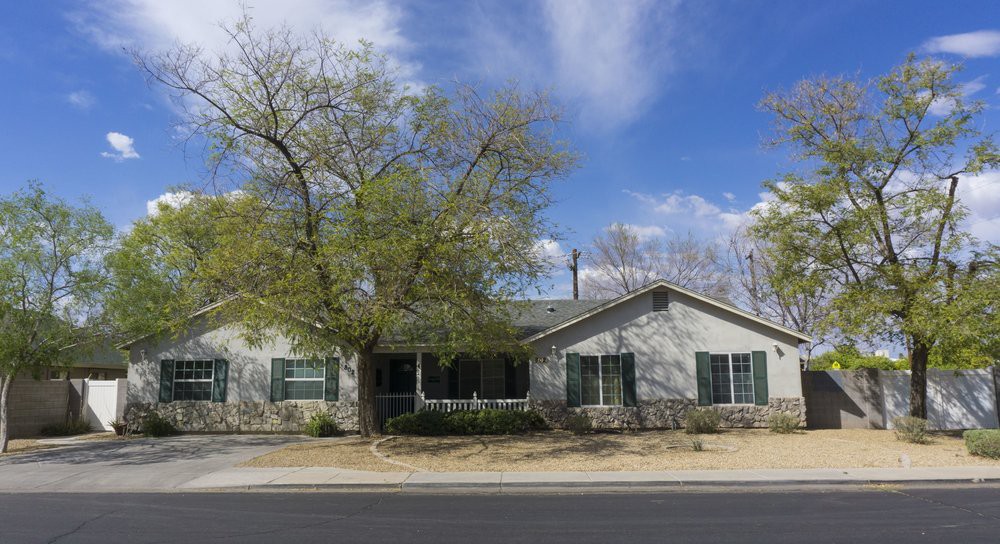 A single-story house with a tree in front, situated along a street under a blue sky with clouds.