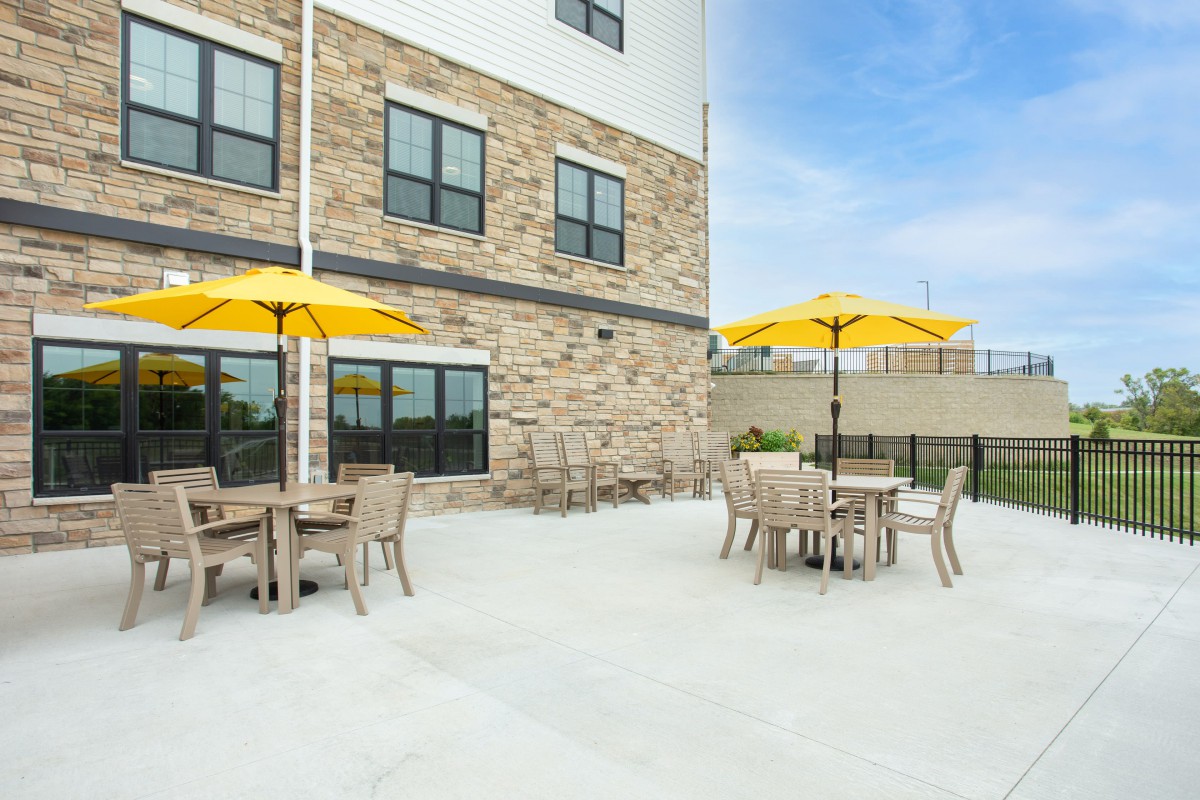 An outdoor patio area with wooden tables and chairs, featuring bright yellow umbrellas against a stone building facade.