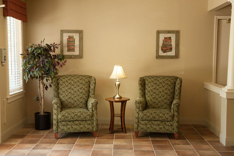 A cozy living room corner with two patterned armchairs flanking a small wooden table topped with a lamp, complemented by framed artwork on the wall and a potted plant in the background.
