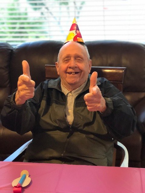 An elderly man wearing a party hat is smiling and pointing, celebrating his birthday.