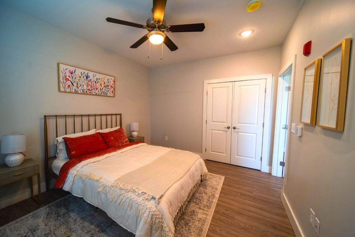 A cozy bedroom featuring a double bed with red pillows, a ceiling fan, and a nightstand with a lamp, all set against hardwood floors.