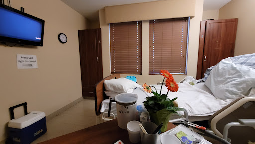 A view of a hospital patient room featuring a bed, medical supplies, and bright window blinds.