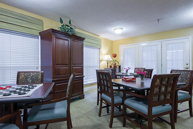 A cozy dining room with a wooden table and chairs, a large cabinet, and light streaming through windows with blinds.