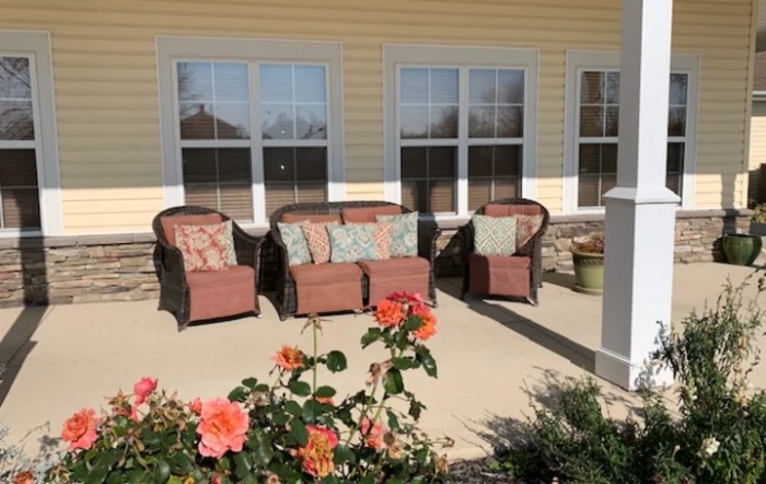 A cozy outdoor patio area with chairs and a sofa, adorned with cushions, surrounded by blooming flowers.