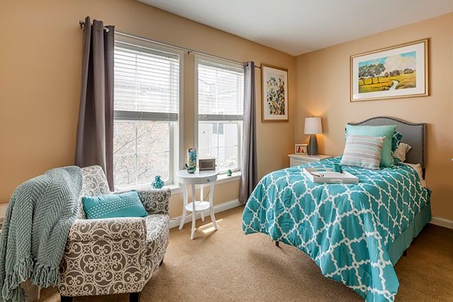 A cozy bedroom with teal and white bedding, a patterned armchair, and large windows with curtains.