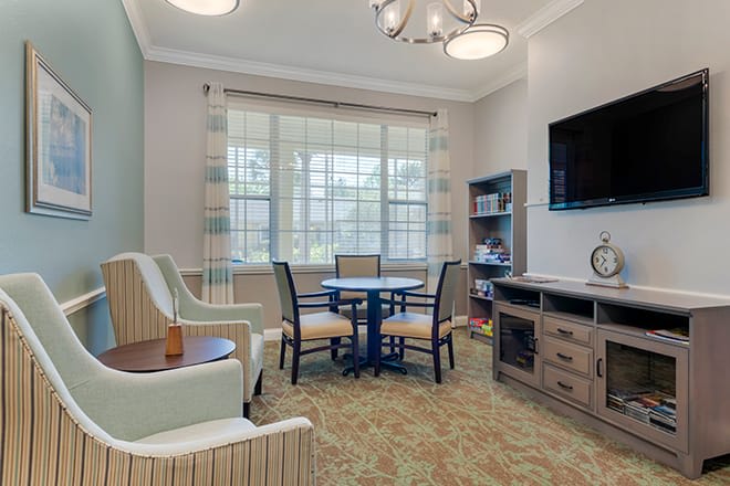 A cozy living room area featuring a round table with chairs, a wall-mounted television, and large windows allowing natural light to illuminate the space.