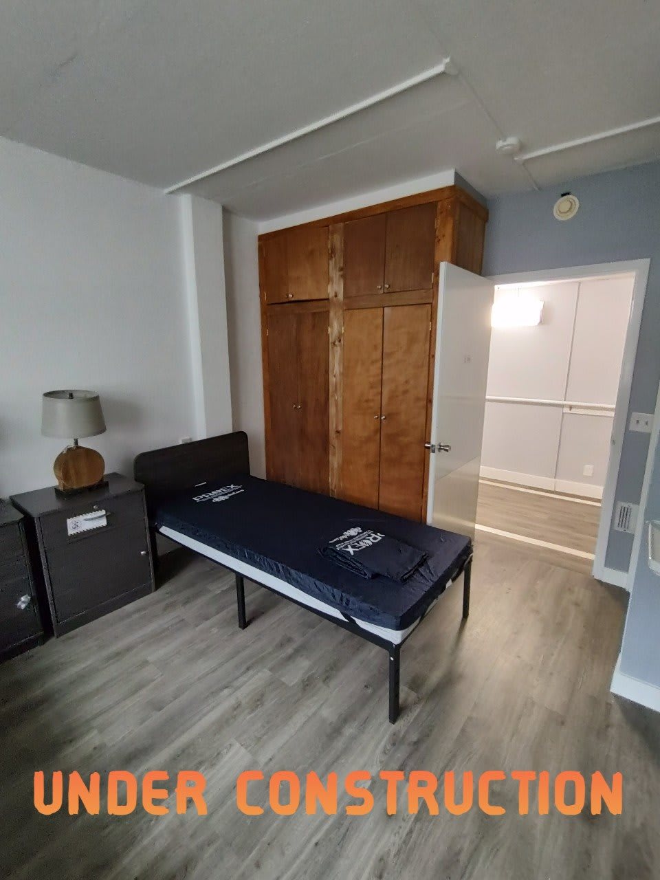 A minimalist bedroom setup with a single bed, wooden wardrobe, and a bedside lamp.