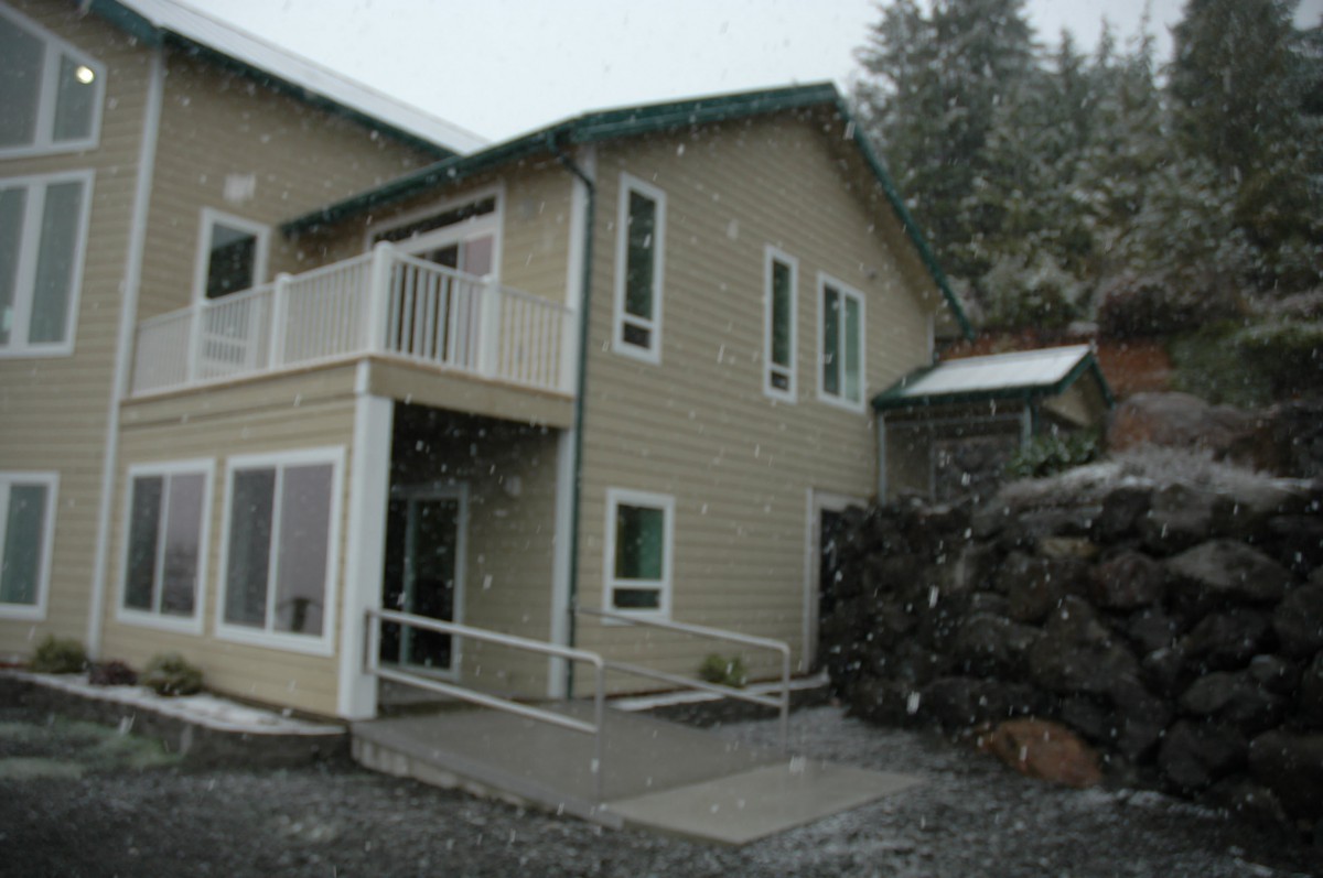 A two-story house with snow falling around it, featuring a balcony and entrance ramp.