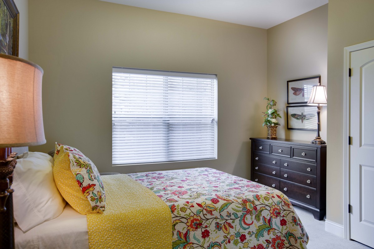 A cozy bedroom with floral bedding, a dark dresser, and natural light coming through a large window.