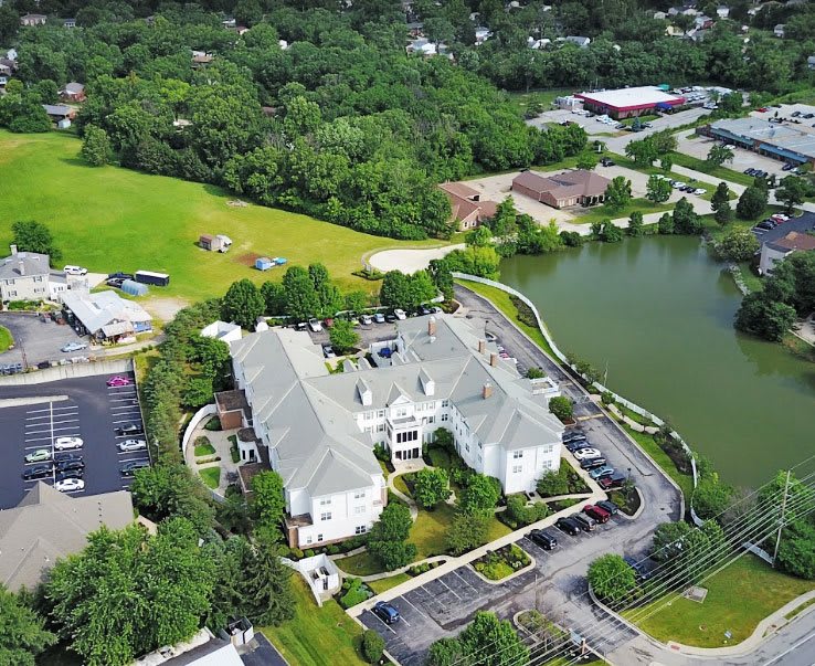 An aerial view of a large building situated next to a lake, surrounded by greenery and parking areas.