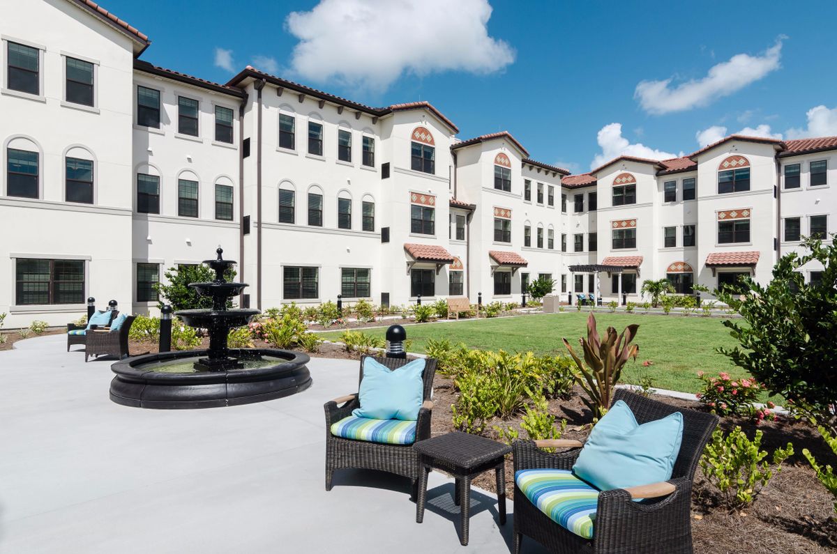 A modern building with outdoor seating, a fountain, and landscaped gardens under a blue sky.