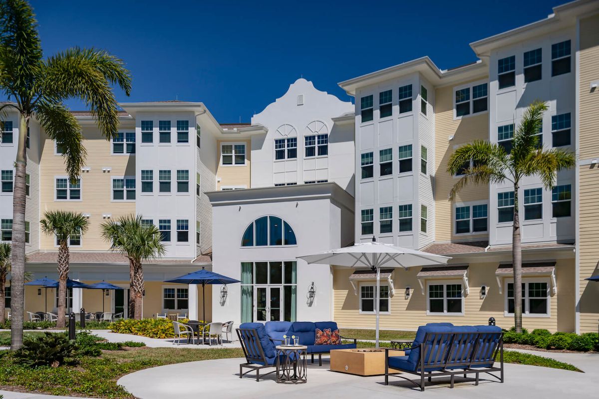 A modern residential building with outdoor seating and palm trees under a clear blue sky.