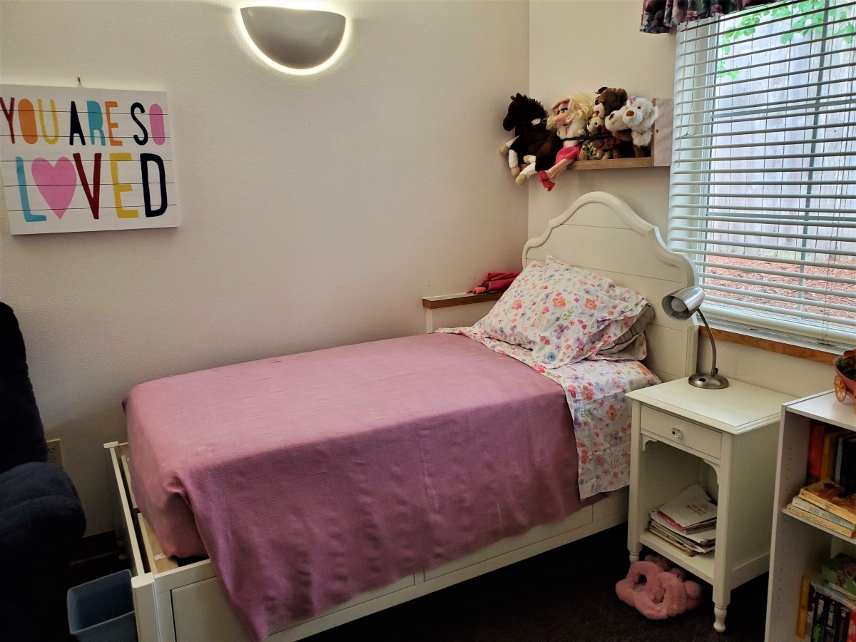 A cozy child's bedroom with a single bed, purple bedspread, stuffed animals, and a nightstand with a lamp.