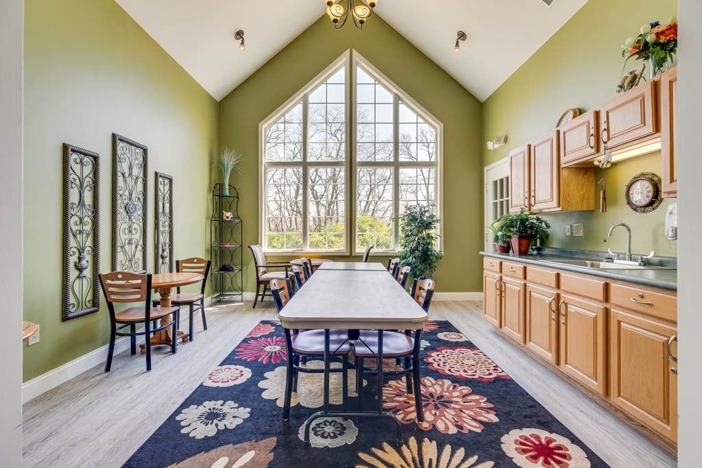 A spacious dining room with a high ceiling and large windows, featuring green walls, wooden furniture, and a decorative rug.