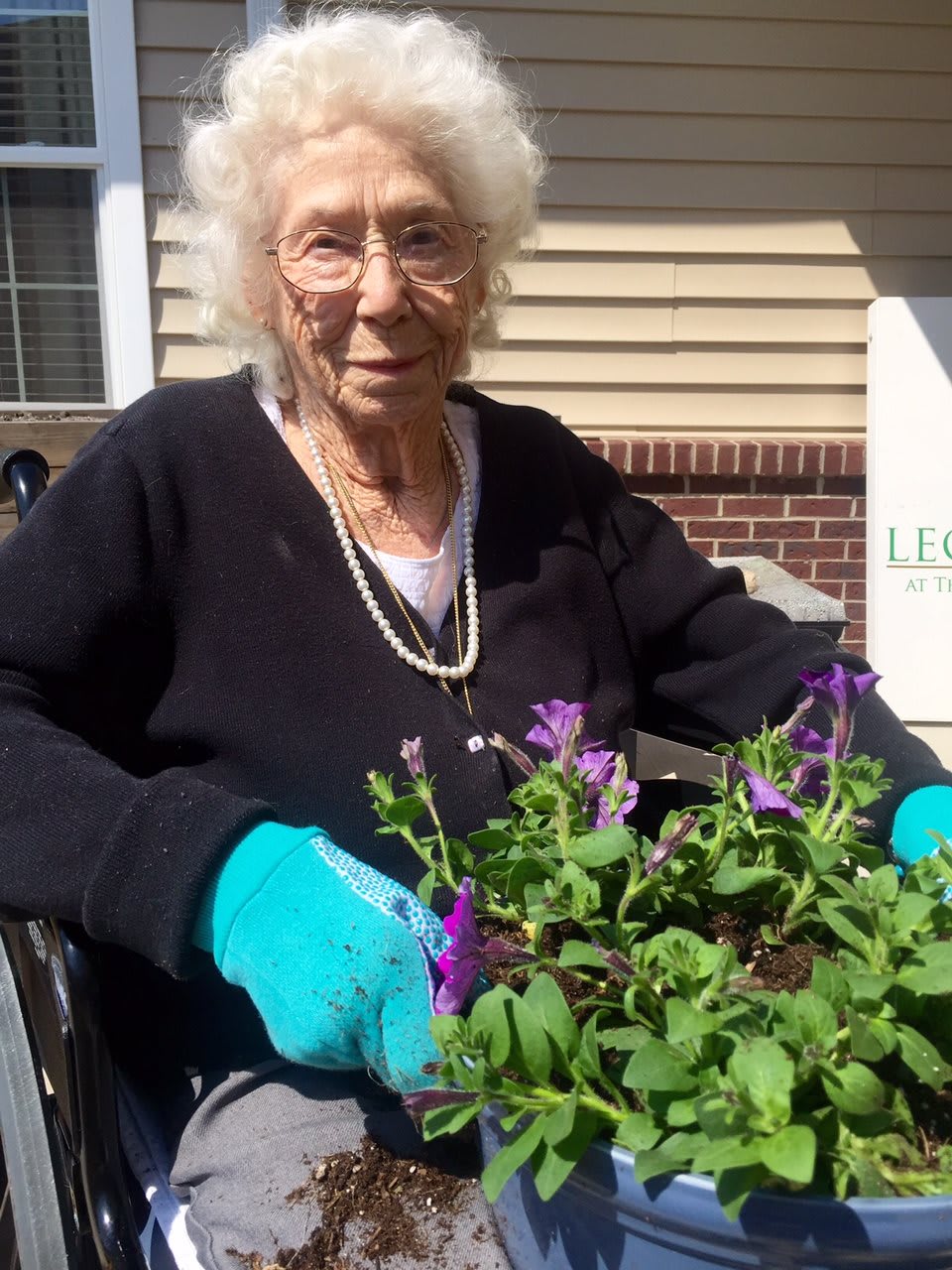 An elderly woman enjoys gardening on a sunny day, tending to her flowers while wearing gloves.