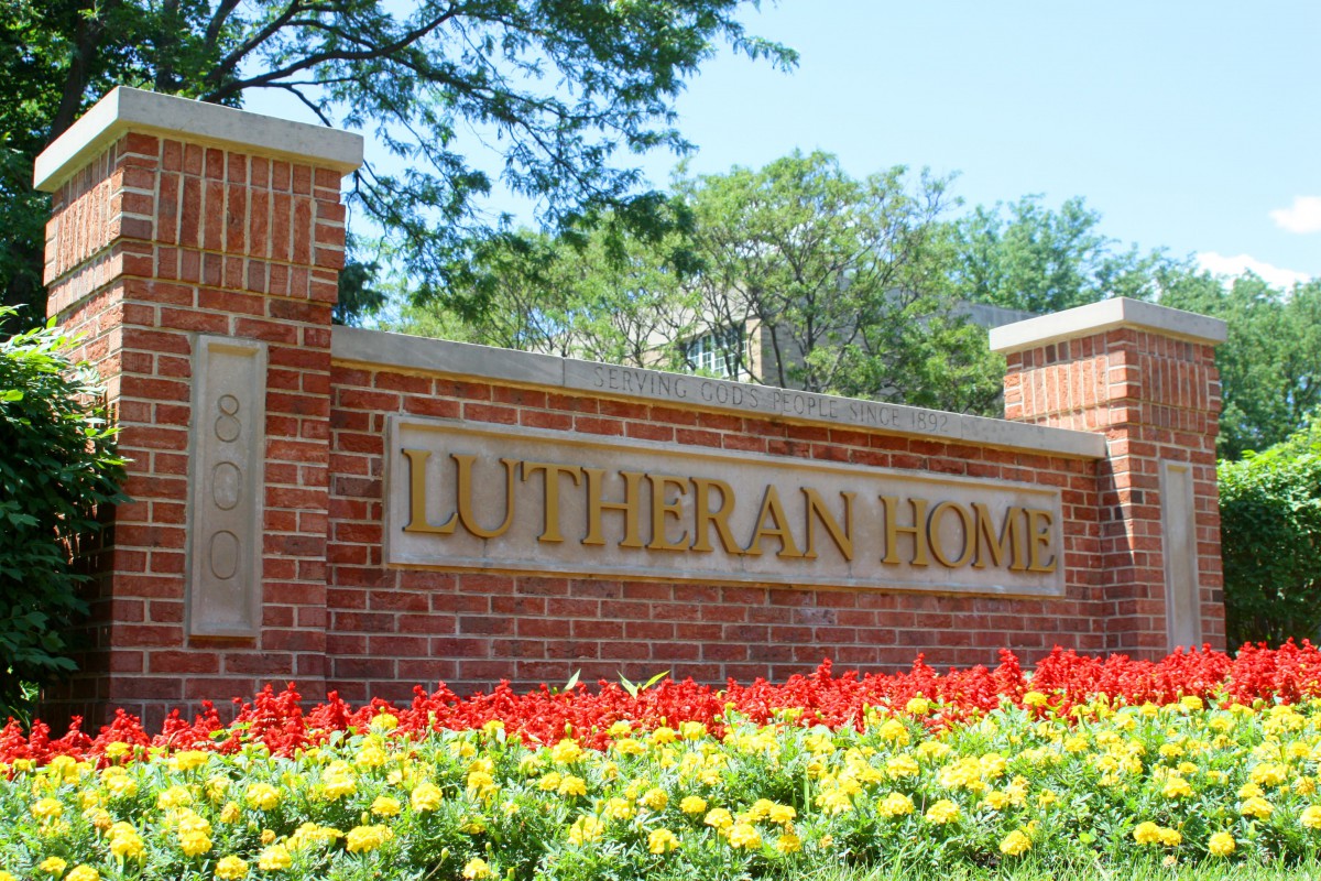 A sign for Lutheran Home on a brick wall surrounded by colorful flowers.