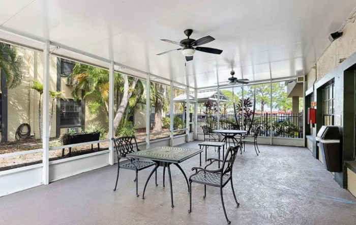 An enclosed outdoor patio with tables, chairs, and ceiling fans, overlooking a lush garden.