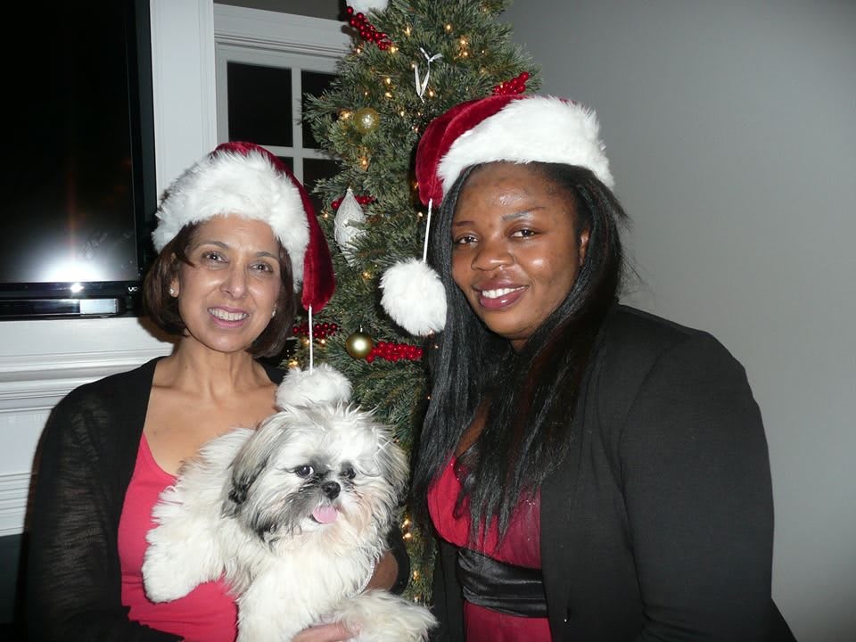 Two women wearing Santa hats celebrate Christmas with a fluffy dog in front of a decorated Christmas tree.