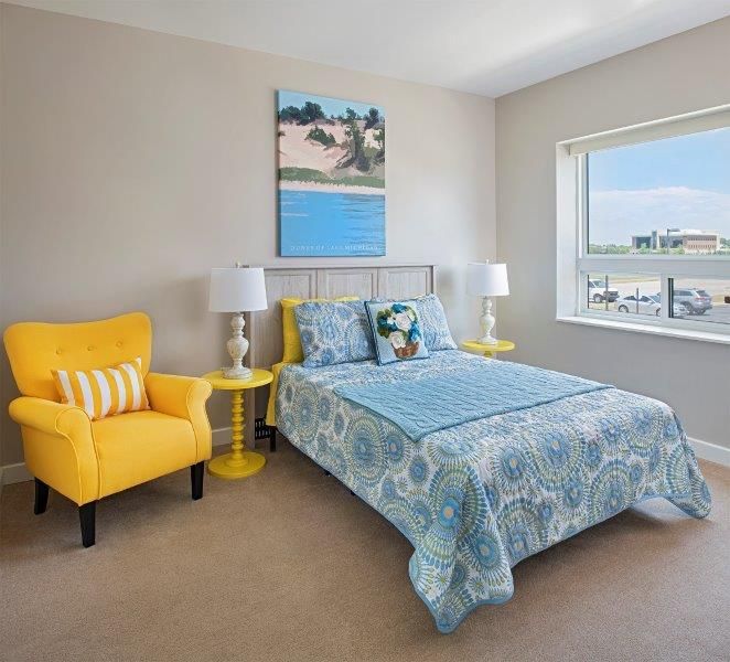 A brightly lit bedroom featuring a blue bedspread, a yellow chair, and a large window with natural light.