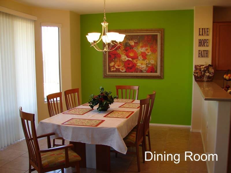 A cozy dining room with a table set for meals, surrounded by wooden chairs and illuminated by a chandelier. A vibrant painting decorates the green accent wall.