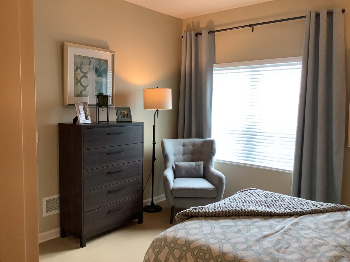 A cozy bedroom with a modern design featuring a dark dresser, an armchair, and a floor lamp by the window.