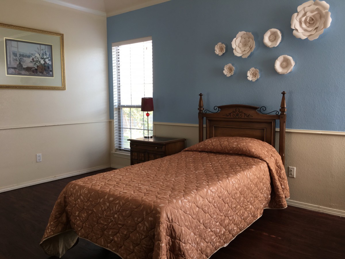 A cozy bedroom features a single bed with an orange-brown quilt, wooden furniture, and decorative wall art on a blue accent wall.