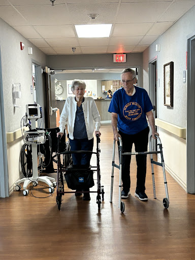 An elderly couple walking together with the help of walkers in a hospital corridor.