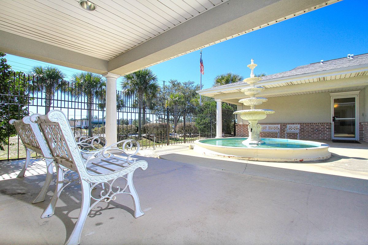 A sunny patio with a white bench and a fountain, surrounded by palm trees.