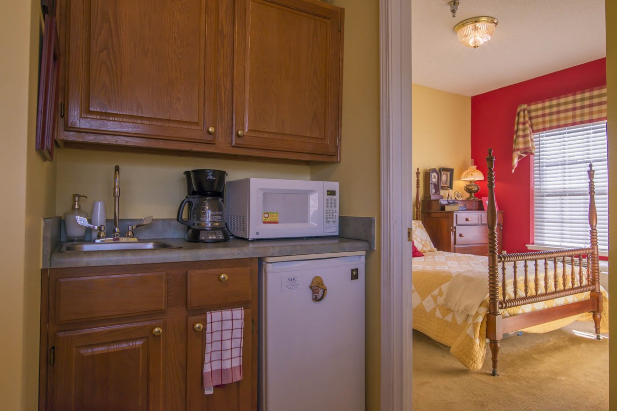 A small kitchenette with a microwave, coffee maker, and mini fridge next to a cozy bedroom.
