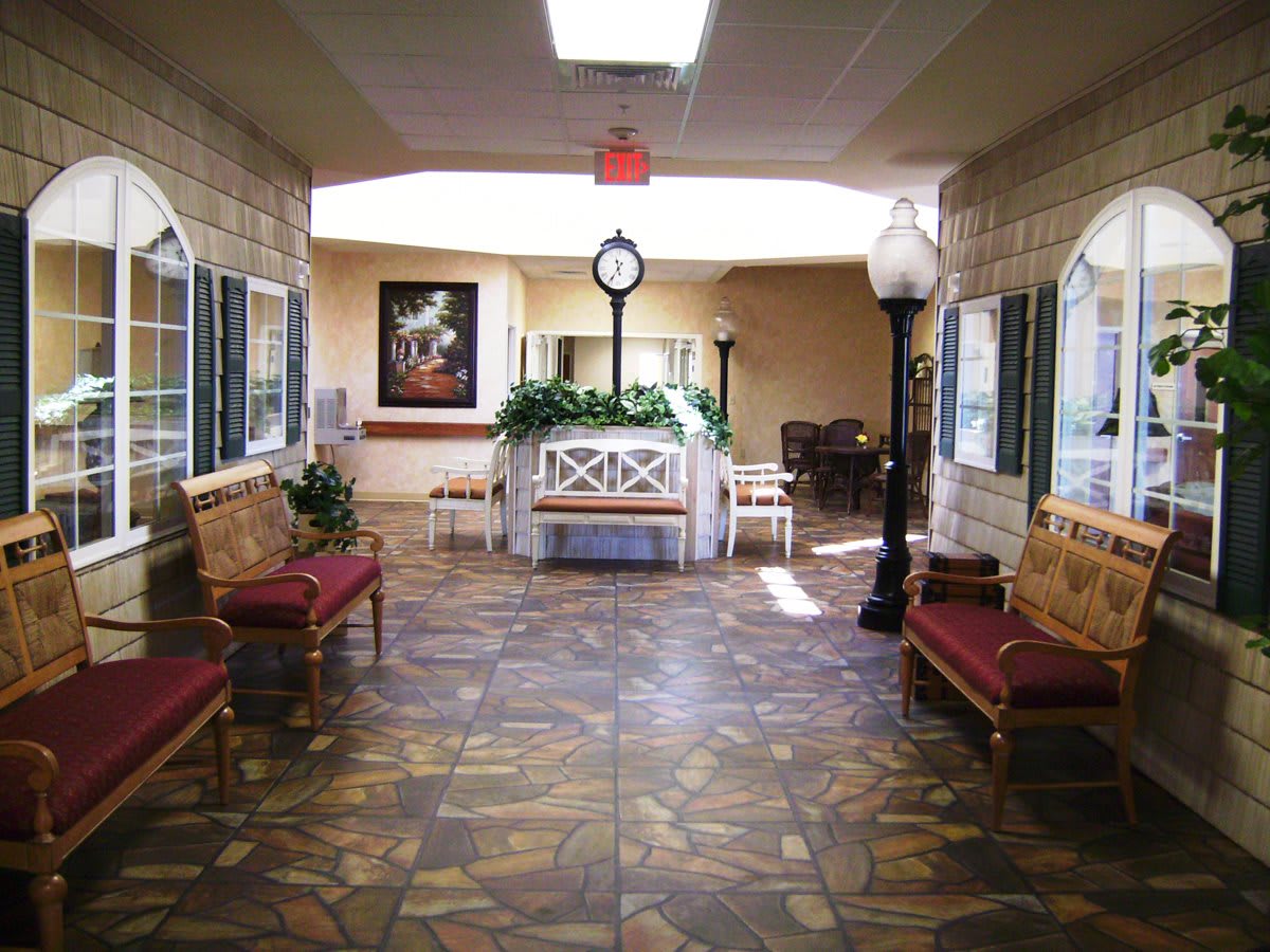 A spacious lobby area featuring decorative flooring, benches, artwork on the walls, and a central clock surrounded by plants.