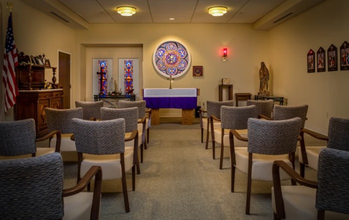 A quiet, intimate chapel with seating and decorative stained glass windows facing a central altar.