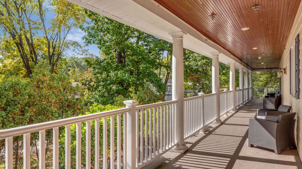 A spacious porch with white columns and railing overlooking lush green trees.