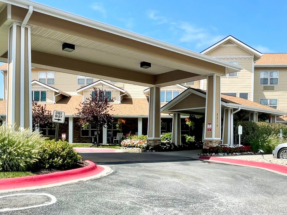 The image shows a building entrance with a covered driveway and landscaped surroundings.